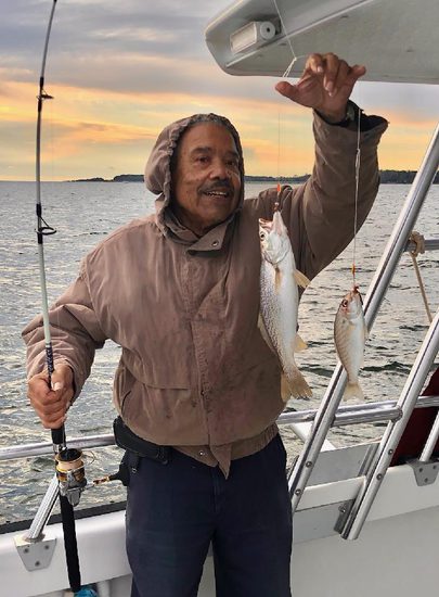A man stands on a boat showing off two fish he just caught.